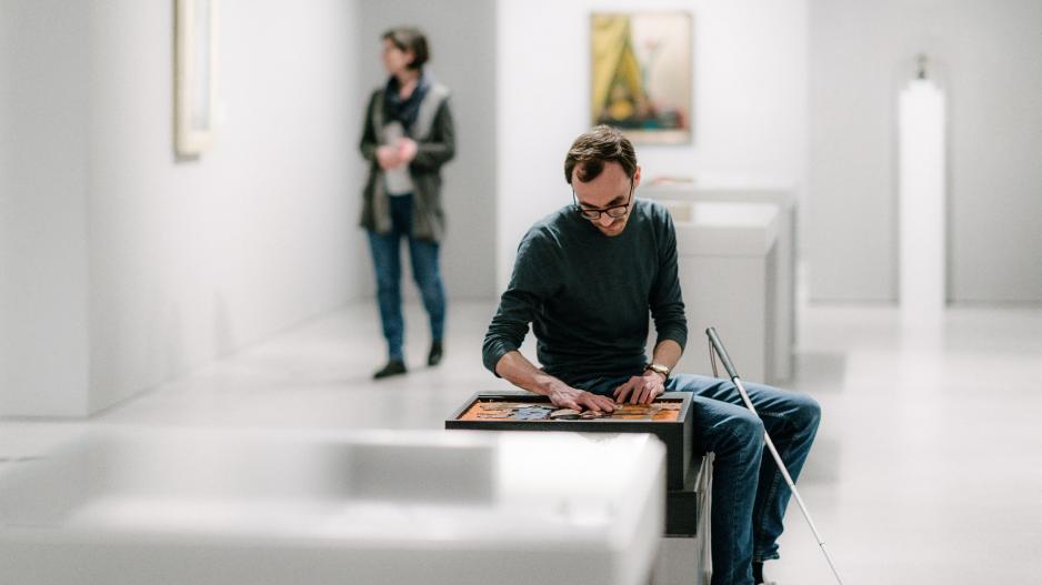 Kunst zum Anfassen in der Berlinischen Galerie. Andreas Krüger sitzt in einem Ausstellungsraum, im Hintergrund sind Bilder aufgehängt, vor einem der Bilder steht eine Besucherin. Vor Andreas Krüger befindet sich ein Tastmodell, das er mit den Händen ertastet, ein Langstock lehnt an seinem Knie.