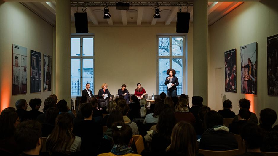Podiumsdiskussion bei der Jahrestagung des LMB 2019. Murat Akan (erster von links) Blick von hinten auf einen vollen Saal. Auf dem Podium sitzen vier Referent*innen, eine Moderatorin mit einem Mikro in der Hand steht daneben.