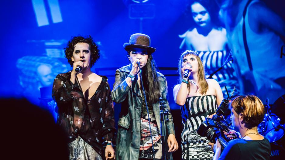 Hieu Pham in der Inszenierung "Der Ring" beim Popkulturfestival Hieu Pham steht mit zwei Kolleg*innen auf der Bühne in der Inszenierung "Der Ring". Die drei singen gemeinsam. Sie trägt eine Melone und einen grünen Ledermantel. Ihr Gesicht ist weiß geschminkt. Sie hält in der rechten Hand ein Mikrofon.